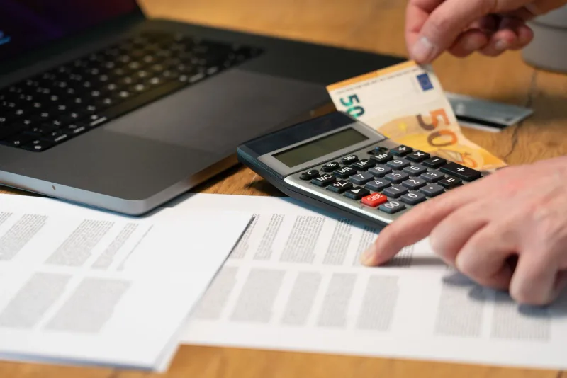 Hands checking paperwork with a calculator, euro banknotes and laptop.